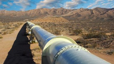 Long white industrial pipeline stretches across a dry desert landscape toward rocky hills under a blue sky.