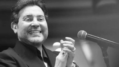 Smiling man in a dark suit claps beside a microphone on stage.