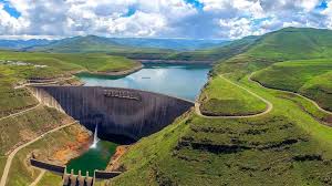 images 43 | MCG24 | موقع إلكتروني مغربي شامل متجدد على مدار الساعة Dam spanning a curved valley with a wide spillway, turquoise reservoir, and emerald green hills under a cloudy sky.