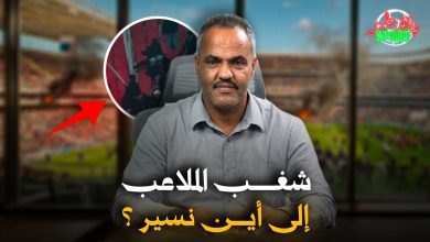 Man in a gray shirt sits at a desk in a studio with a blurred sports stadium background; a circular inset shows a crowd, plus a red arrow and Arabic headline text overlaid.
