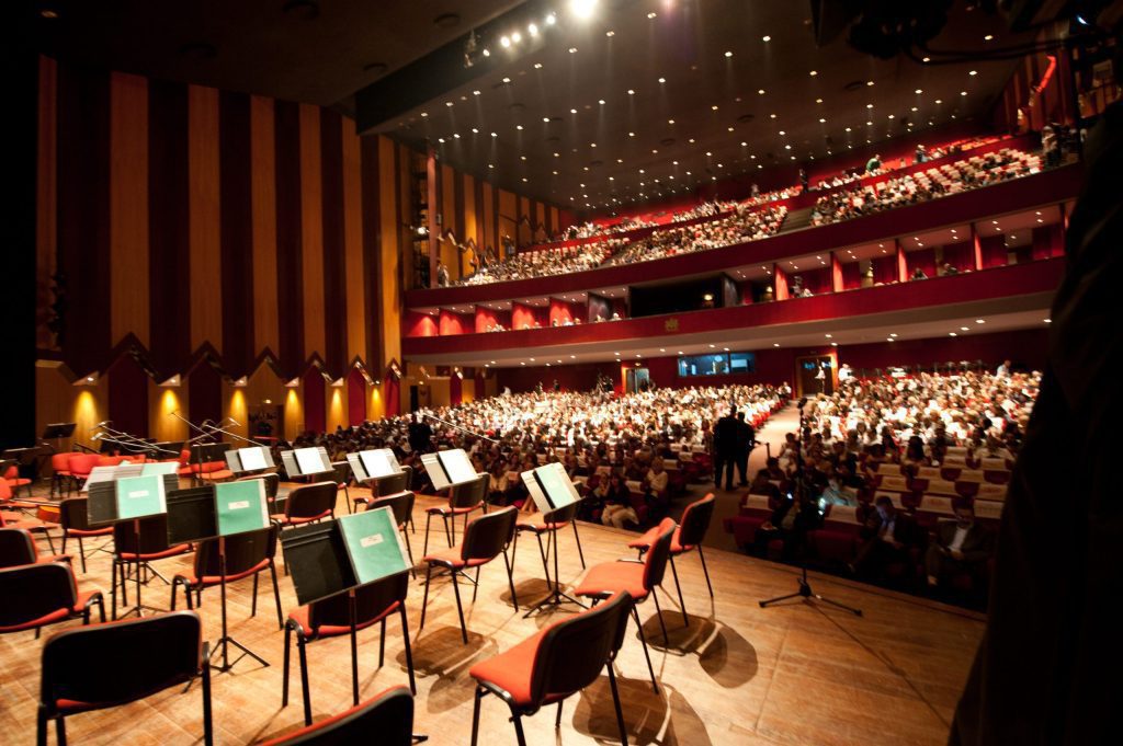 الرباط تحتضن الدورة 23 للمسابقة الوطنية للموسيقى Concert hall interior with a stage full of empty chairs and music stands in the foreground and a large audience seated in red seats on multiple balconies above.