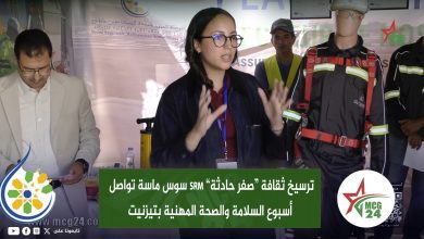 Woman with glasses speaks at a conference, flanked by a man in a suit and a rescue worker in safety gear beside her, with an Arabic banner behind.