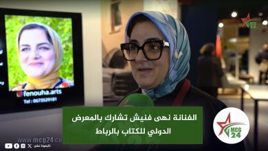 Woman in a blue patterned hijab and black glasses being interviewed at a media event, holding a green microphone with MC G24 logo visible in the background.
