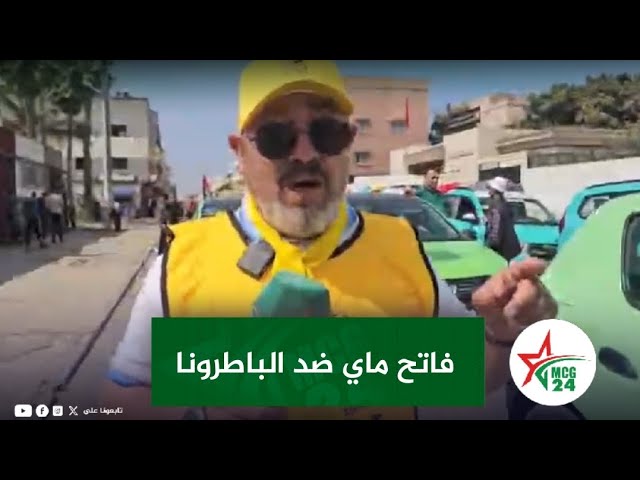 Reporter in a yellow vest and cap speaks into a green mic on a city street; Arabic banner on screen and cars in the background.