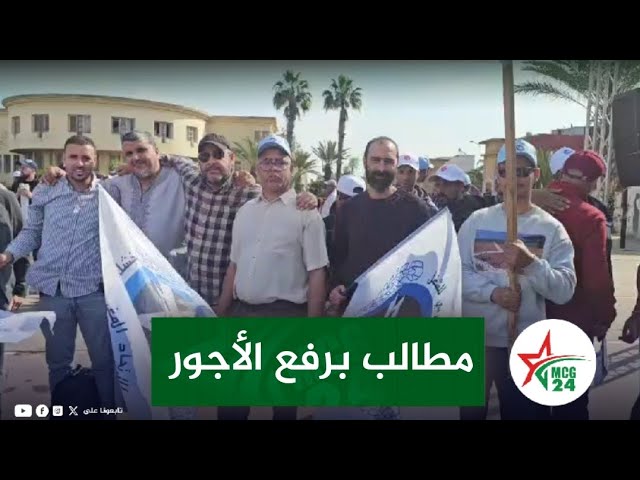 Group of men standing together in a street protest, holding flags, with a green banner that reads 'مطالب برفع الأجور' (Demands to raise wages).