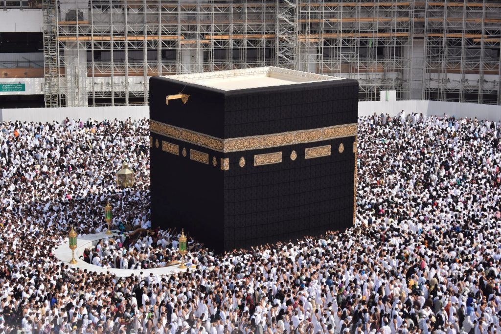 unsplash-image-IAwnp88Fz8Y | MCG24 | موقع إلكتروني مغربي شامل متجدد على مدار الساعة Black Kaaba with gold-embroidered bands surrounded by a dense crowd of pilgrims in white ihram garments; construction scaffolding in the background.