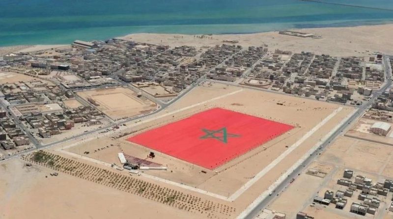 vis_0119202622370481 2 | MCG24 | موقع إلكتروني مغربي شامل متجدد على مدار الساعة Aerial view of a coastal desert town with a large red square on the ground, bearing a green Moroccan pentagram emblem in the center.