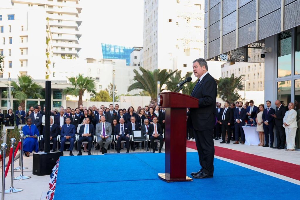 vis_0501202606354426 | MCG24 | موقع إلكتروني مغربي شامل متجدد على مدار الساعة Man in a suit speaks at a podium on a blue-carpeted outdoor stage as a formal ceremony unfolds, with a crowd behind him.