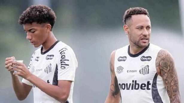 Two male soccer players in Santos FC training shirts on a practice field; left player checks a phone, right looks ahead.