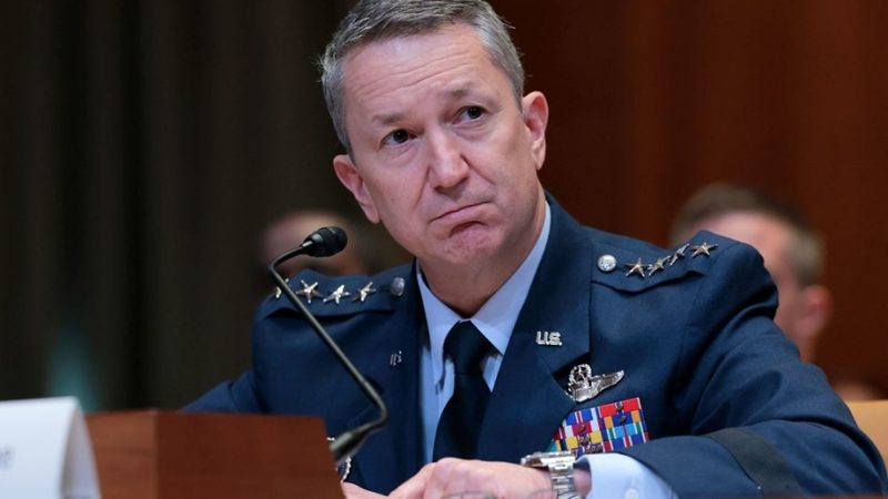 Air Force general in a blue dress uniform sits at a panel with a microphone, looking to the side as he speaks or listens.