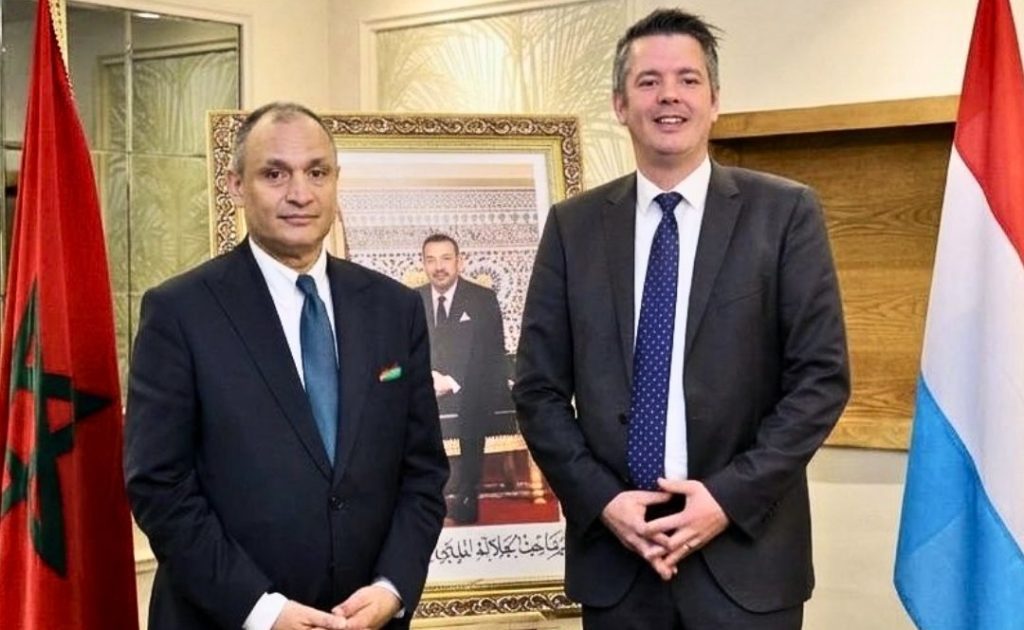 vis_0507202616360742 | MCG24 | موقع إلكتروني مغربي شامل متجدد على مدار الساعة Two men in dark suits pose for a formal photo in an office, with a framed portrait and flags on either side behind them.