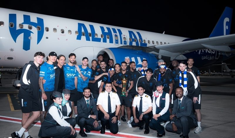 vis_0509202600293706 | MCG24 | موقع إلكتروني مغربي شامل متجدد على مدار الساعة Group of airline crew and athletes posing with a trophy beside a blue-and-white airplane at night on the tarmac.