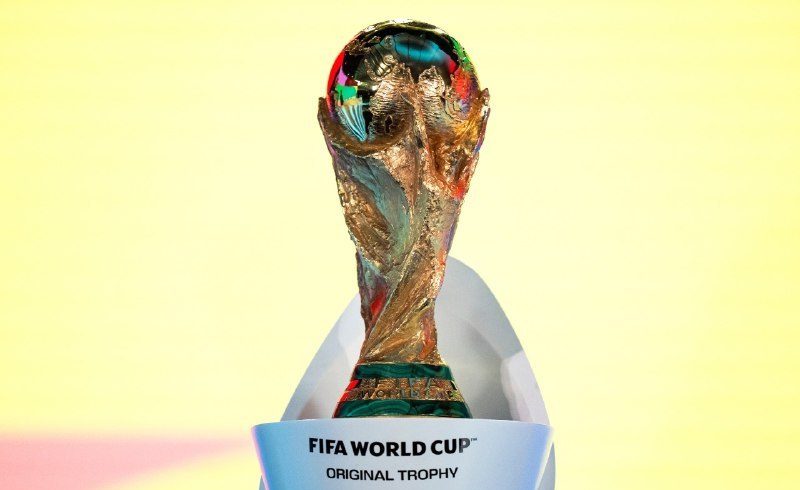 vis_0509202609230774 | MCG24 | موقع إلكتروني مغربي شامل متجدد على مدار الساعة Gold FIFA World Cup original trophy with colorful reflections, set against a pale yellow background.