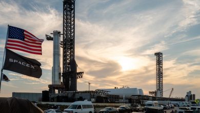 SpaceX launch pad with a rocket, waving American flag and SpaceX banner, cars parked in the foreground at sunset with a bright sky.