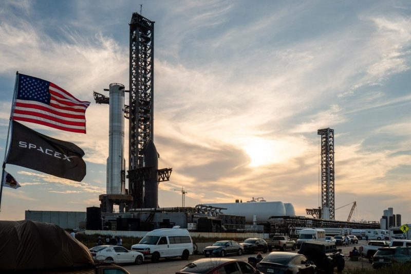 SpaceX launch pad with a rocket, waving American flag and SpaceX banner, cars parked in the foreground at sunset with a bright sky.