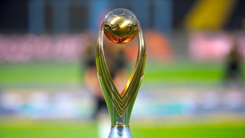 vis_0522202622133390 | MCG24 | موقع إلكتروني مغربي شامل متجدد على مدار الساعة Gold sports trophy with a round golden ball atop a curved green base on a blurred stadium field in the background, symbolizing victory.