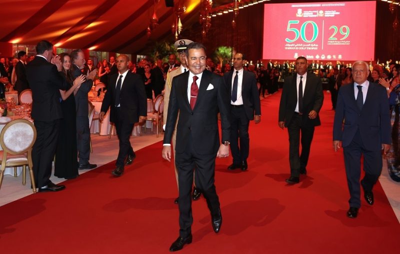 vis_0522202623003833 | MCG24 | موقع إلكتروني مغربي شامل متجدد على مدار الساعة Smiling man in a dark suit and maroon tie walks the red carpet at a formal gala, with guests and a large red screen behind.