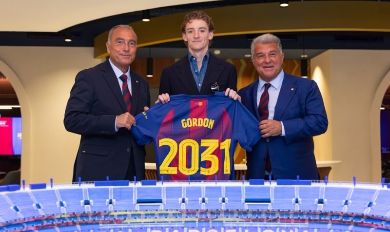 Three men in suits present a blue jersey labeled Gordon and 2031, standing behind a scale model stadium.