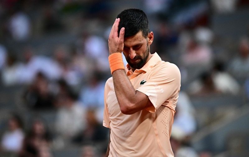 vis_0529202622082802 | MCG24 | موقع إلكتروني مغربي شامل متجدد على مدار الساعة Male tennis player in peach shirt touching his forehead with his right hand on the court, orange wristband on his wrist, blurred crowd in background.
