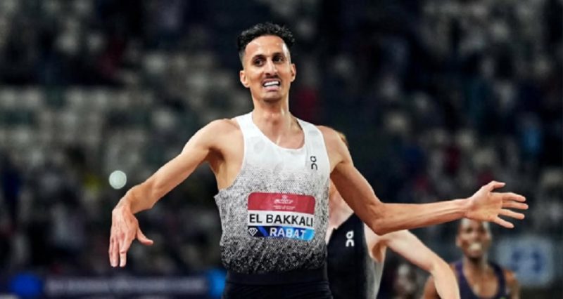vis_0531202621223713 | MCG24 | موقع إلكتروني مغربي شامل متجدد على مدار الساعة Male track athlete in a white gradient singlet with a red El Bakkali bib, arms outstretched in celebration on a stadium track.