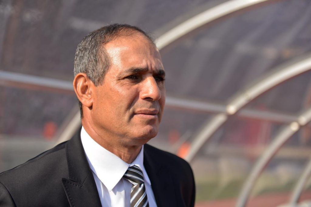 2456688 | MCG24 | موقع إلكتروني مغربي شامل متجدد على مدار الساعة Middle-aged man in a black suit and striped tie stands outdoors, looking to the right near a curved shelter or dugout.