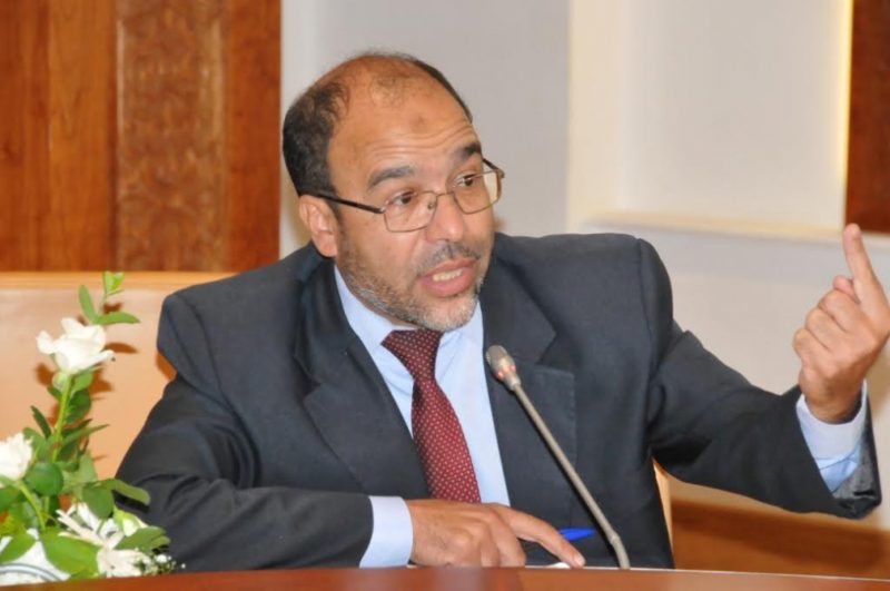 أحمد-صدقي | MCG24 | موقع إلكتروني مغربي شامل متجدد على مدار الساعة Man in a dark suit and red tie speaking into a microphone at a panel, gesturing with his right hand while seated at a table with flowers nearby.