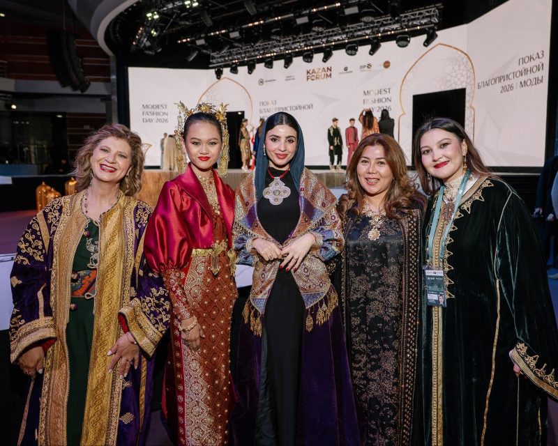 القفطان المغربي في روسيا | MCG24 | موقع إلكتروني مغربي شامل متجدد على مدار الساعة Five women in ornate, traditional embroidered dresses smiling for a photo at a fashion show stage with a Kazan Forum backdrop behind them.