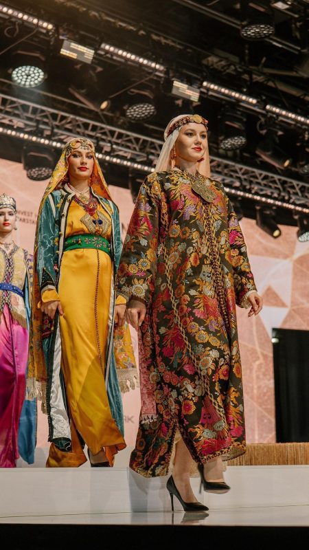 القفطان المغربي في روسيا2 | MCG24 | موقع إلكتروني مغربي شامل متجدد على مدار الساعة Models wearing ornate traditional dresses walk a fashion show runway, vibrant floral embroidery and headpieces visible under stage lights.