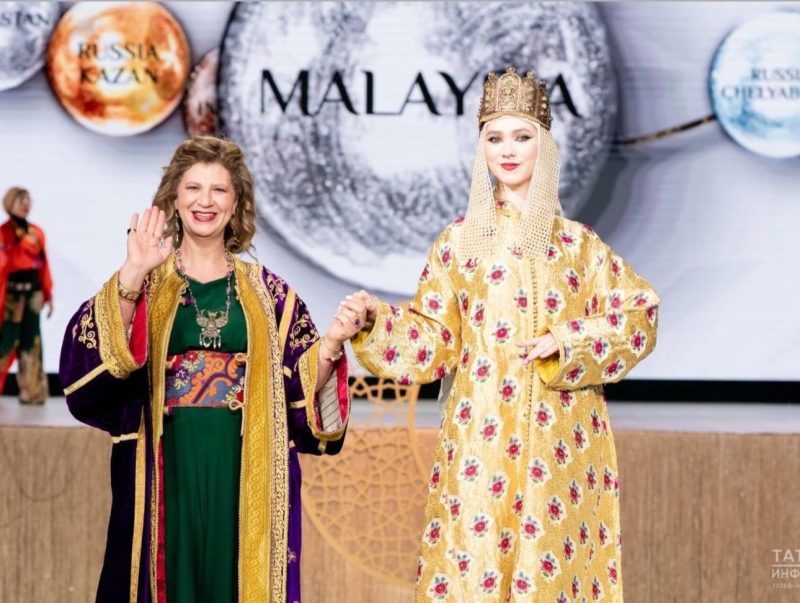 القفطان المغربي في روسيا5 | MCG24 | موقع إلكتروني مغربي شامل متجدد على مدار الساعة Two women in ornate traditional Malaysian outfits on a stage; one waves while the other, in a gold floral gown with a crown, holds her hand.