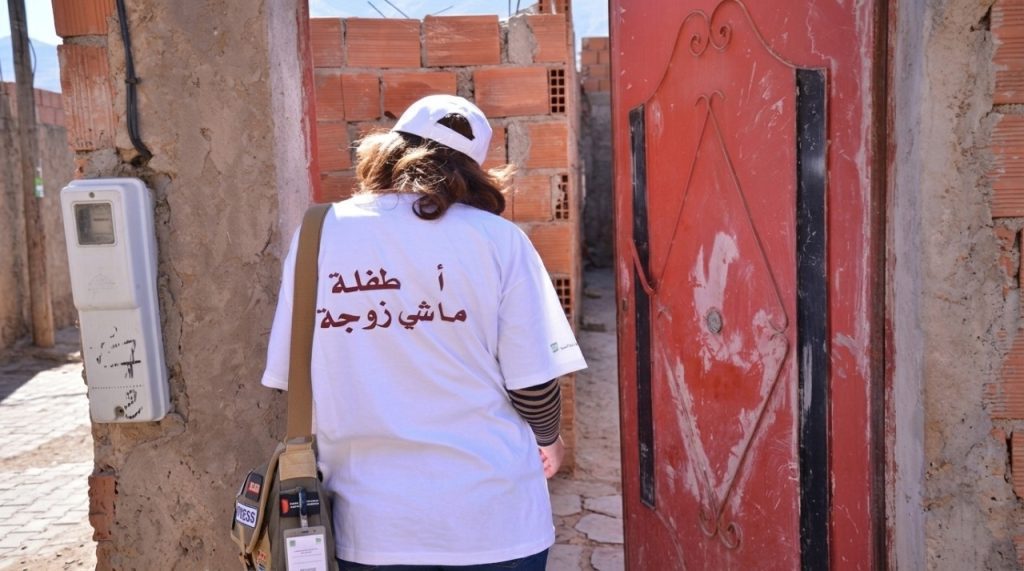 جمعيات ضد تزويج القاصرات | MCG24 | موقع إلكتروني مغربي شامل متجدد على مدار الساعة Person wearing a white T‑shirt with Arabic text on the back, carrying a tan shoulder bag and press badge, walking through a brick-walled entry beside a red door.