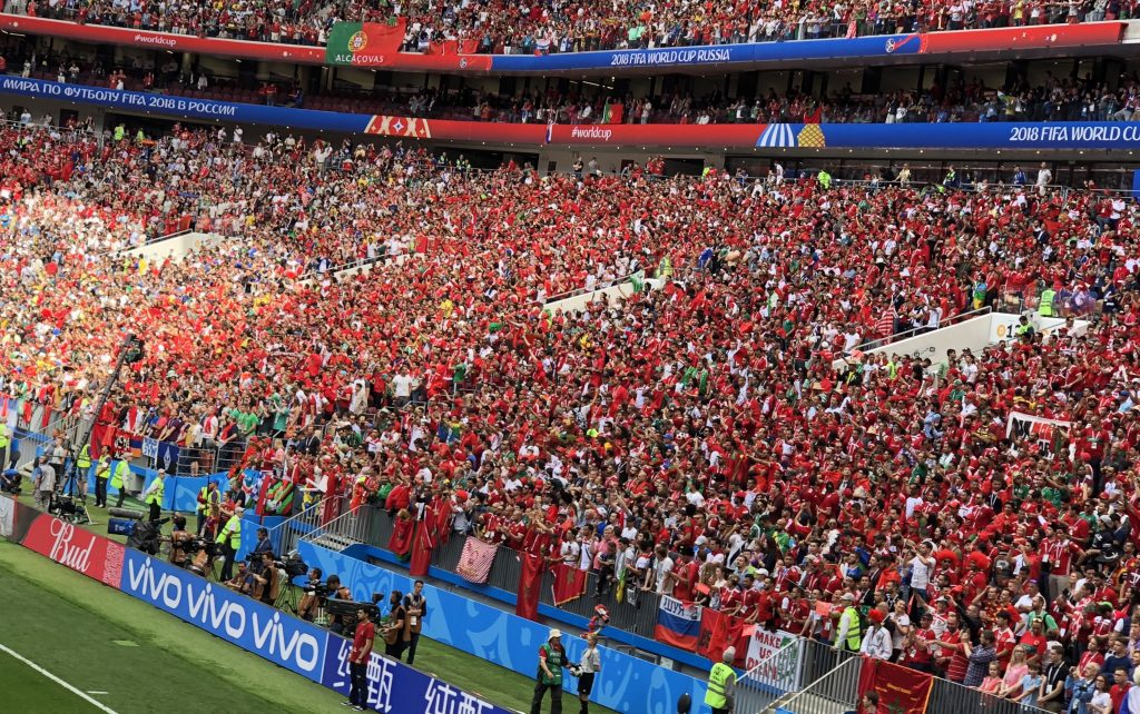 Crowd of football fans wearing red shirts filling a stadium during a 2018 FIFA World Cup match with banners overhead reading Russia/Russia 2018 World Cup material]
