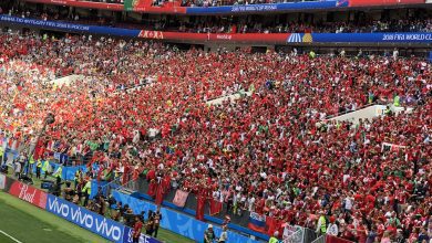 Crowd of football fans wearing red shirts filling a stadium during a 2018 FIFA World Cup match with banners overhead reading Russia/Russia 2018 World Cup material]