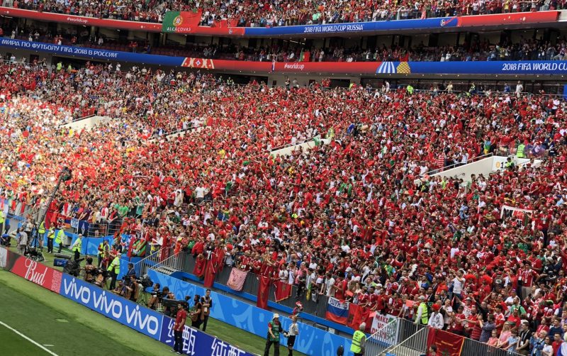 Crowd of football fans wearing red shirts filling a stadium during a 2018 FIFA World Cup match with banners overhead reading Russia/Russia 2018 World Cup material]