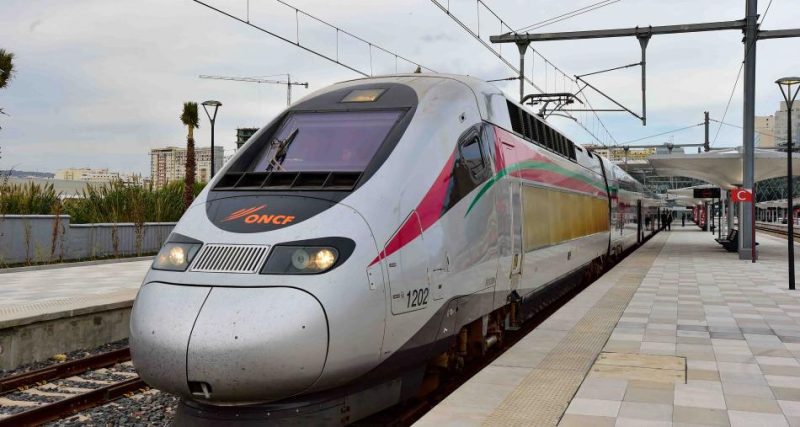 ALBOURAK - TGV MAROCAINE LE 16-11-2018 163 1 | MCG24 | موقع إلكتروني مغربي شامل متجدد على مدار الساعة High-speed train at a platform, with overhead electric lines and a modern white exterior featuring pink and green stripes and the ONCF logo on the front.