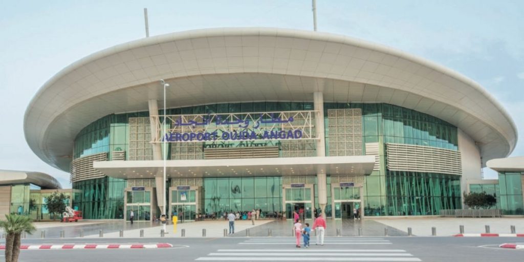 Aeroport-Oujda-Angad | MCG24 | موقع إلكتروني مغربي شامل متجدد على مدار الساعة Oujda Angads Airport terminal with a curved white roof, glass walls, and people walking toward the entrance under a clear sky.