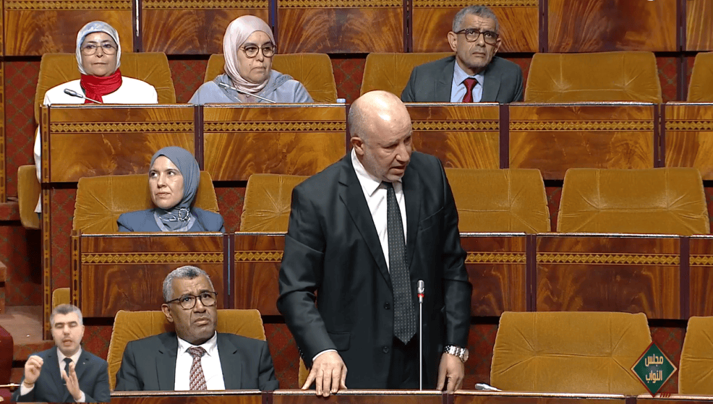 Capture-decran-2025-11-03-165713 | MCG24 | موقع إلكتروني مغربي شامل متجدد على مدار الساعة Bald man in a dark suit speaking at a microphone in a wood-paneled parliamentary chamber, with several seated attendees behind him in orange-brown seats.