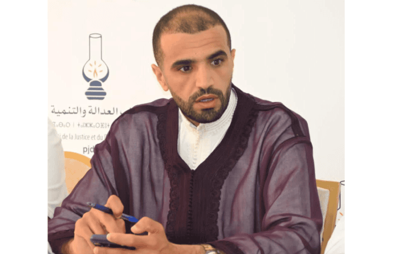 Capture-decran-2026-05-31-142553 | MCG24 | موقع إلكتروني مغربي شامل متجدد على مدار الساعة Man in a maroon traditional robe sits at a meeting, looking to the side while holding a phone and a pen.