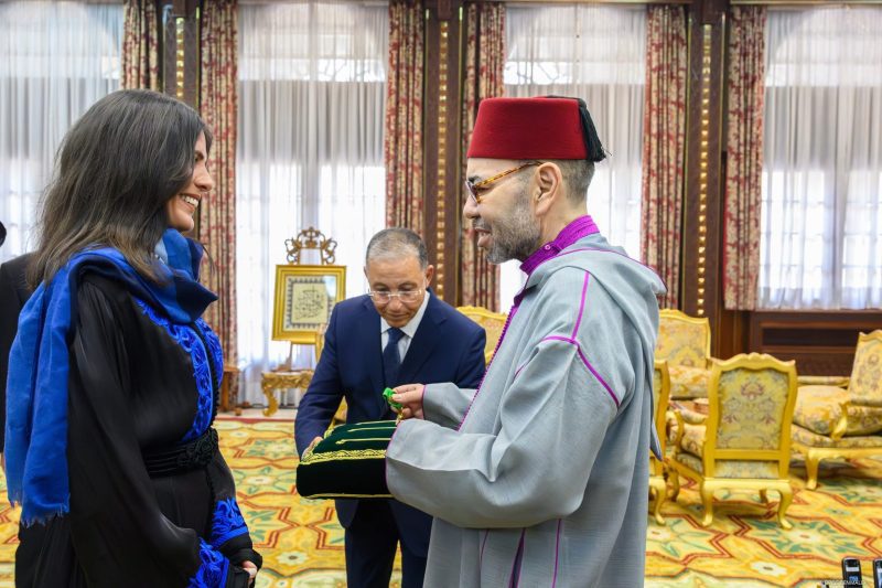Smiling woman in blue and black attire receives a green item from a man in gray ceremonial robes and a red fez during a formal ceremony.