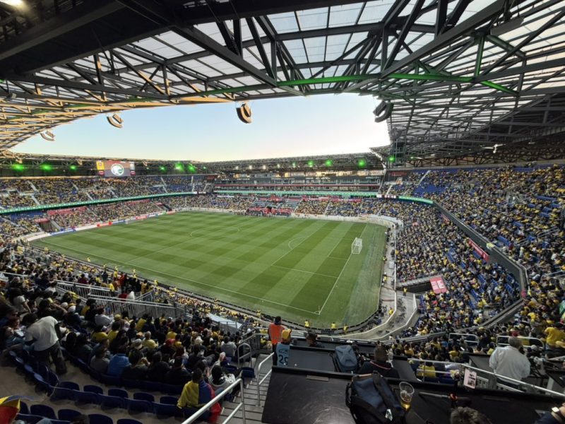 Wide-angle view of a soccer stadium packed with fans, a green field in the center, and a roofed stands above.