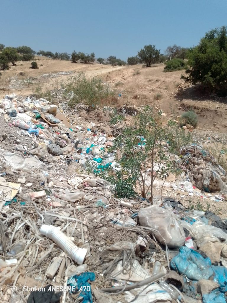 Pile of garbage and plastic waste scattered in a dry, hilly landscape with sparse trees in the distance and a clear blue sky at top.
