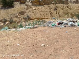 Trash and discarded tires piled at the base of a rocky hillside, with plastic bags and litter scattered on the dirt ground near sparse vegetation