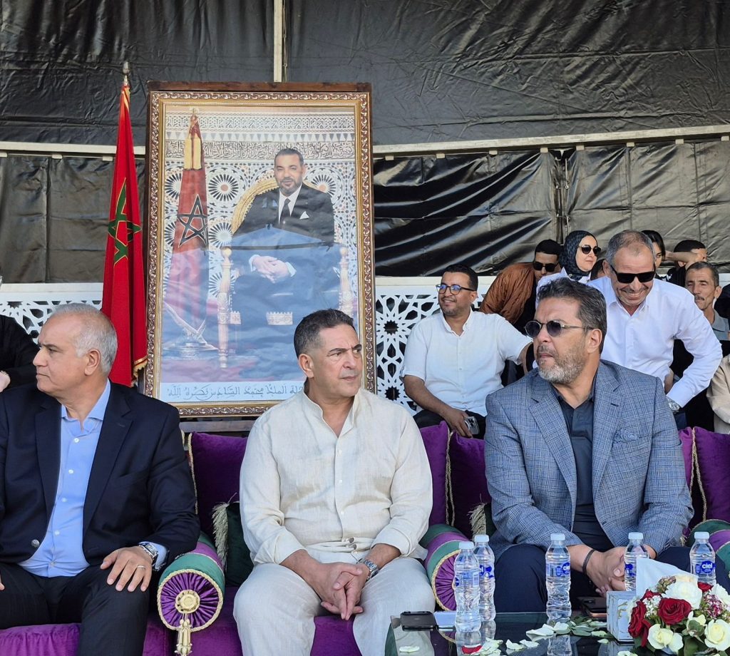 الوالي أمزازي يترأس فعاليات الدورة التاسعة لكرنفال بيلماون بودماون Outdoor ceremony with men in suits seated, next to a large ornate framed portrait and a Moroccan flag behind them.