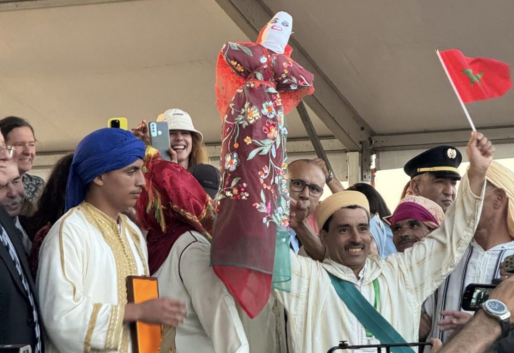 Group of Moroccan performers in traditional dress celebrating, one person holds up a colorful floral shawl-wrapped figure atop a stick amid smiling crowd and a red flag in the background