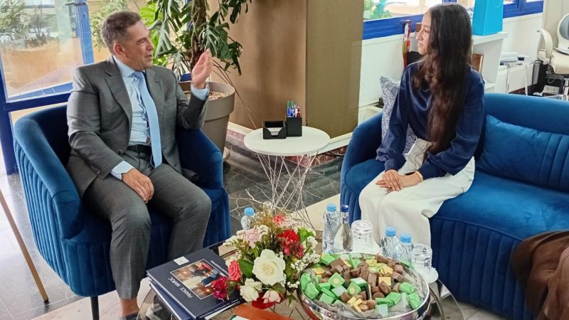Two adults sit on blue velvet couches in a modern lobby, engaged in conversation as the man gestures with his hand.] .