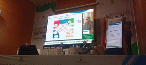Speaker at podium beside a large projection displaying a colorful diagram during a conference event.
