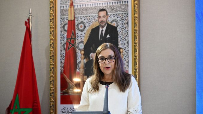 IMG_2682-678x381-1 | MCG24 | موقع إلكتروني مغربي شامل متجدد على مدار الساعة Woman wearing glasses speaks at a podium; Moroccan flag visible to her left and a framed portrait of King Mohammed VI behind her.