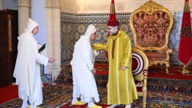 Moroccan king in a gold-yellow caftan greets a guest as two officials in white robes observe, in a richly tiled palace with flags and an ornate throne.