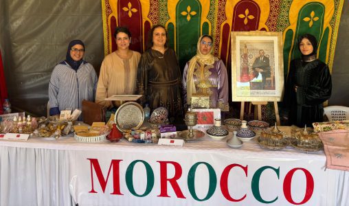 Nouveau-projet216 | MCG24 | موقع إلكتروني مغربي شامل متجدد على مدار الساعة Six women stand behind a Moroccan craft stall displaying metalware, pottery, textiles, and a framed portrait, with a colorful backdrop and a bold 'MOROCCO' banner.