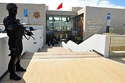Operator__BCIJ__stands_guard_in_the_headquarters_in_the_city_Salé | MCG24 | موقع إلكتروني مغربي شامل متجدد على مدار الساعة Armored security officer stands at attention outside a modern public building entrance with stairs and glass doors behind him.