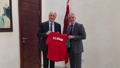 Two men in suits hold a red T-shirt printed with 'M.S. BERRADA' in a formal room.